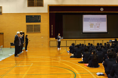 写真：会場の体育館でスクリーンを使用して説明するパートナーと座って聞いている生徒たち